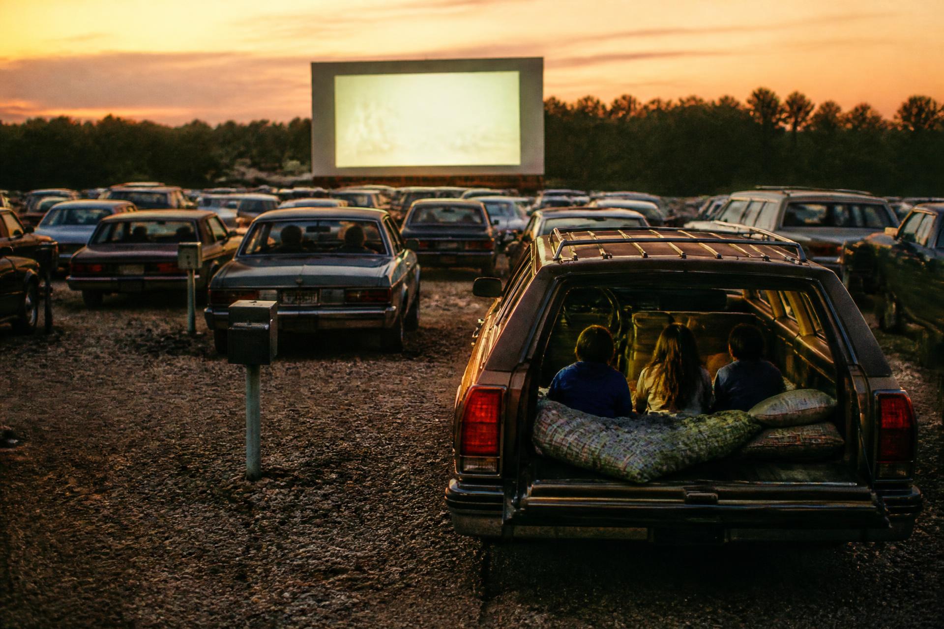 Cars filled with families at an outdoor drive-in theater at dusk in 1987, all facing the screen